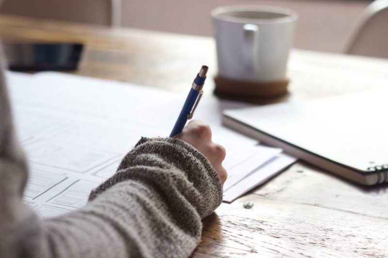 A close up of documents and papers and a hand holding a pen with a coffee cup on the background